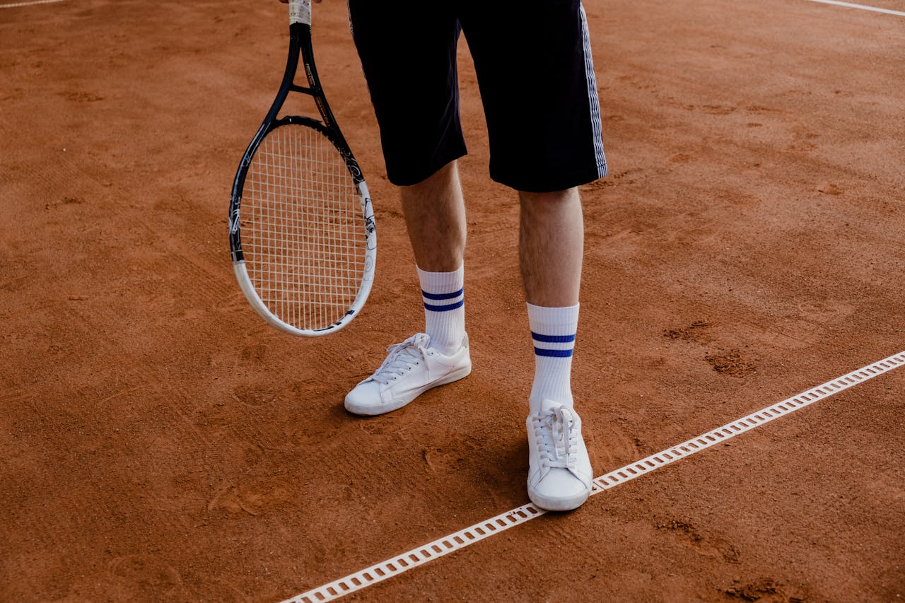 Man standing on a clay tennis court holding a racket, wearing sneakers.